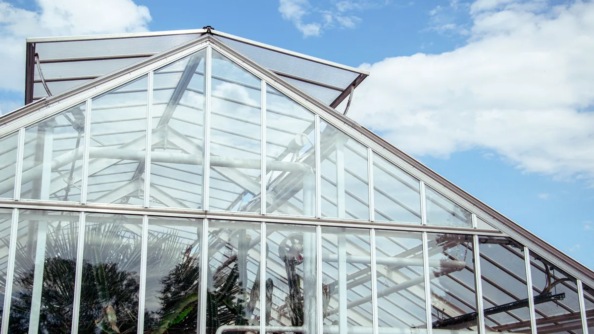 Tejado a dos aguas de un invernadero de cristal con estructura metálica, con plantas visibles a través del cristal y un cielo azul con nubes.